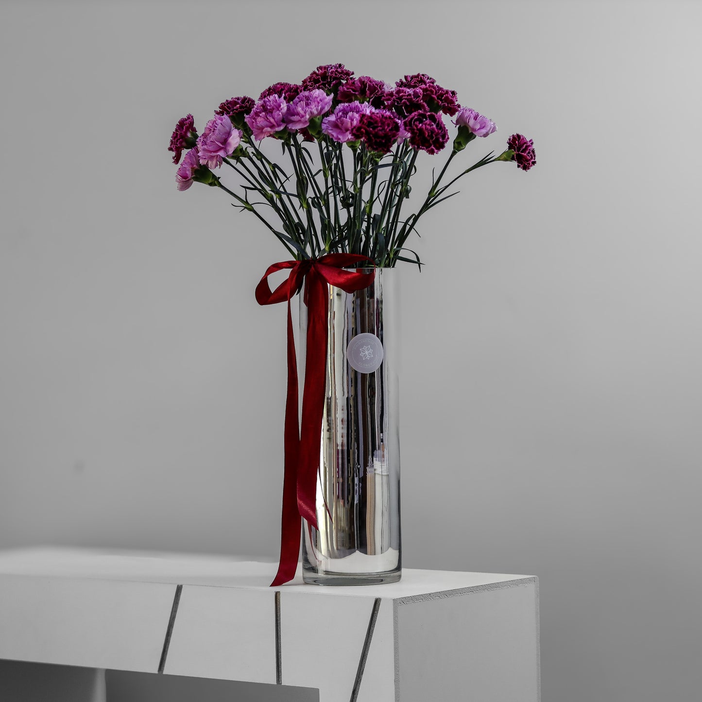 Tall mirror-silver cylinder vase with magenta and plum carnations tied with a red ribbon