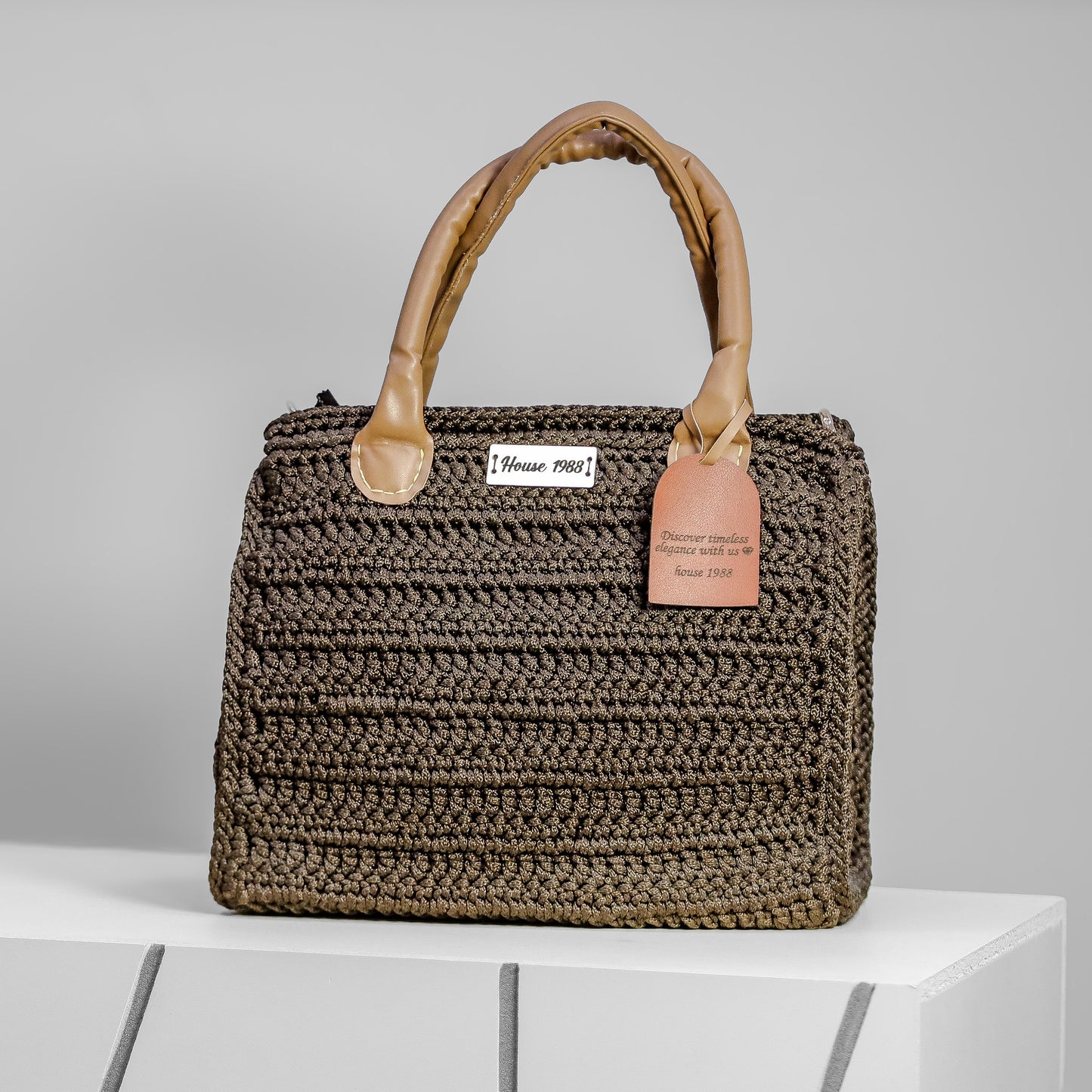 House 1988 handmade crochet handbag in dark taupe with padded tan handles and logo tag on front