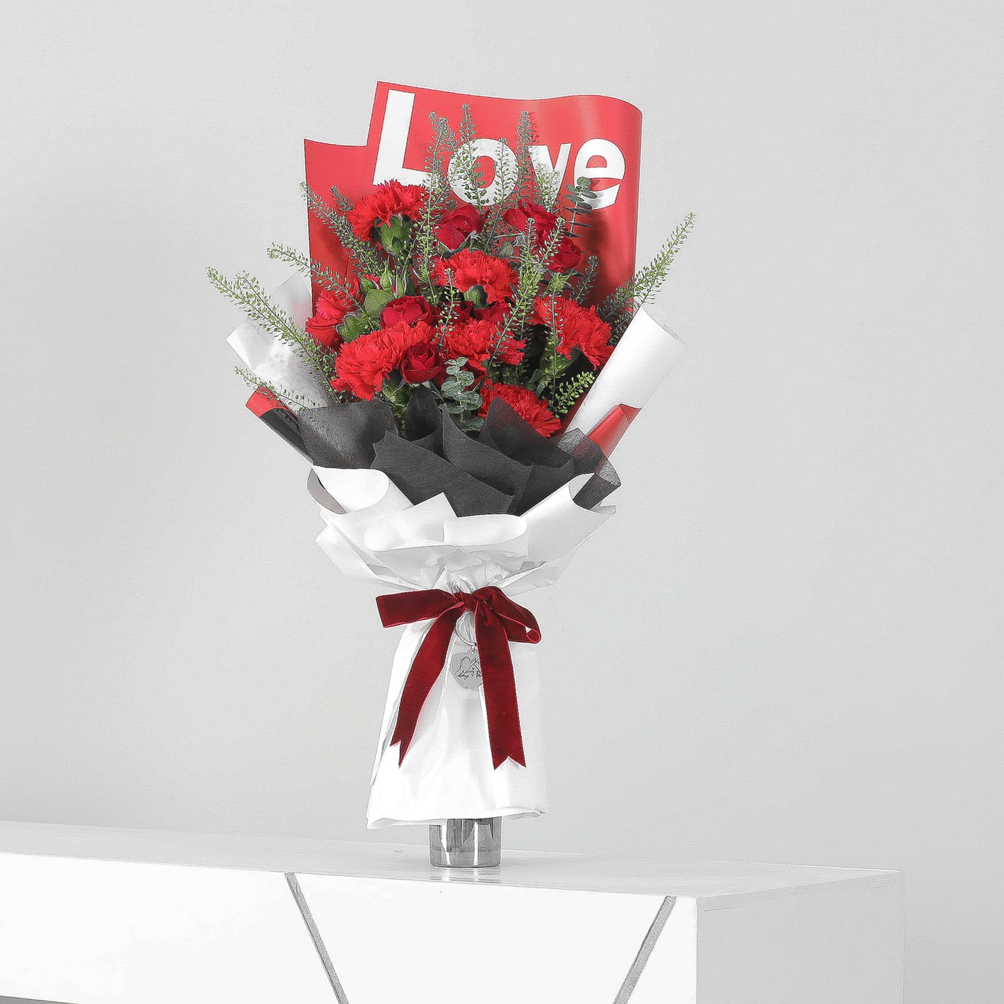 Luxurious red carnation bouquet with greenery in white and black wrapping with “Love” script and red velvet ribbon
